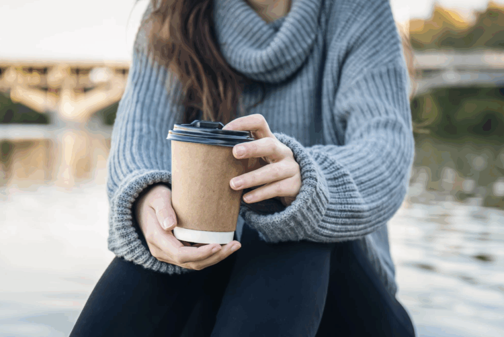A person in a cozy gray sweater holding a hot coffee cup by a serene lake.