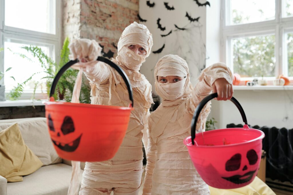 2 kids dressed as Mummies for Halloween holding out candy boo baskets
