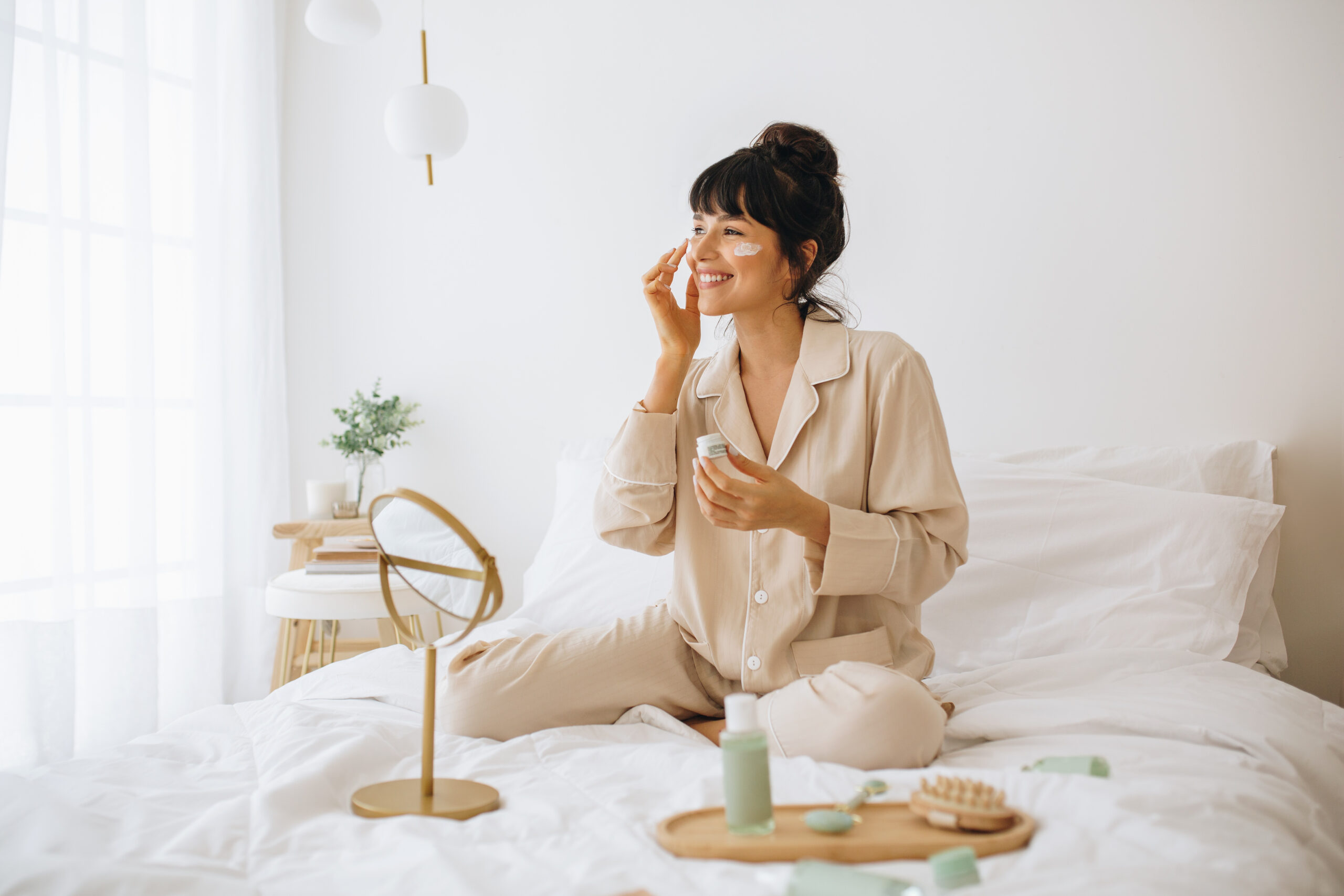 A woman smiles and sits on a bed. She is applying face cream.