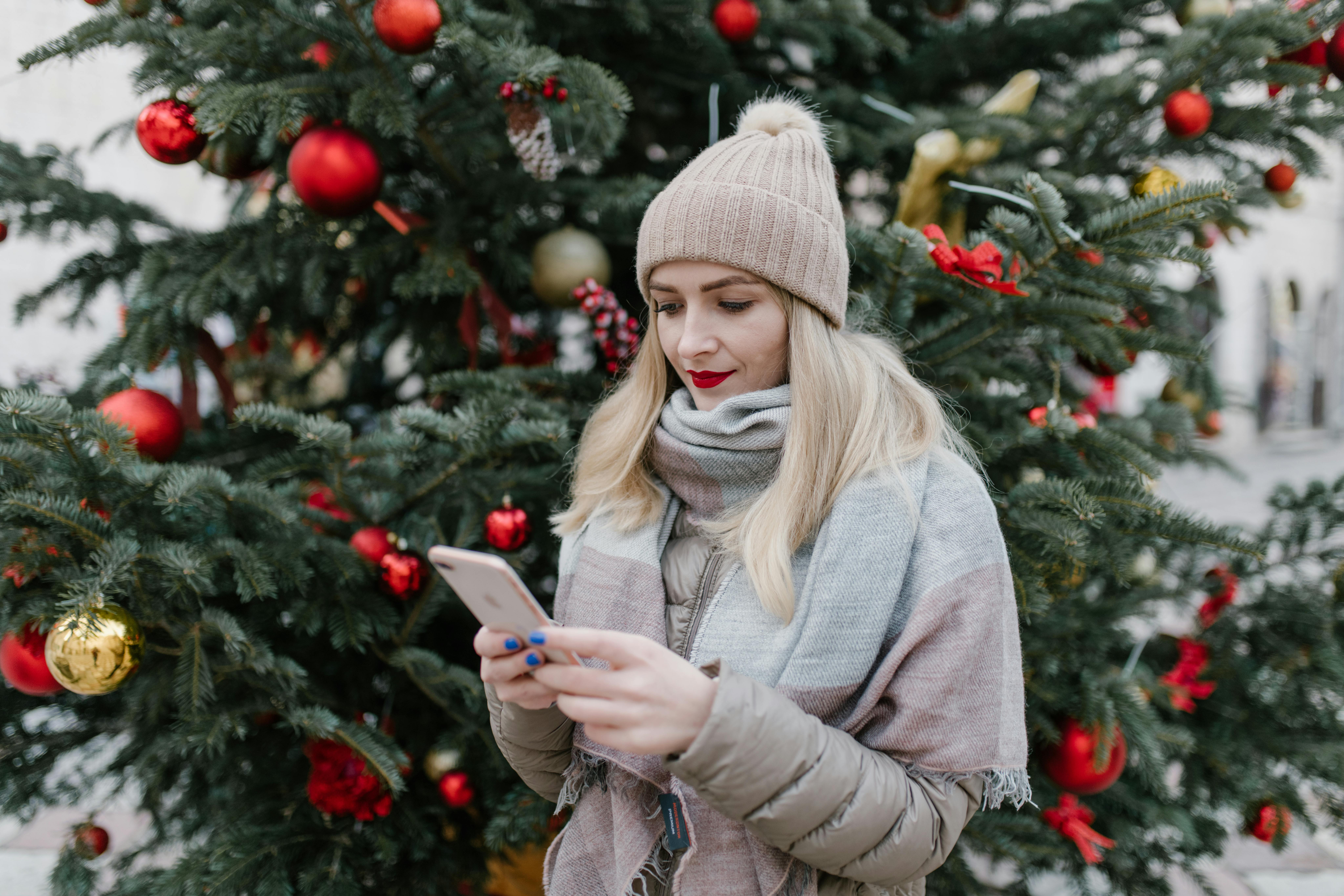 Woman Shopping for Christmas Gifts on phone in front of Decorated Christmas tree with red ornaments.