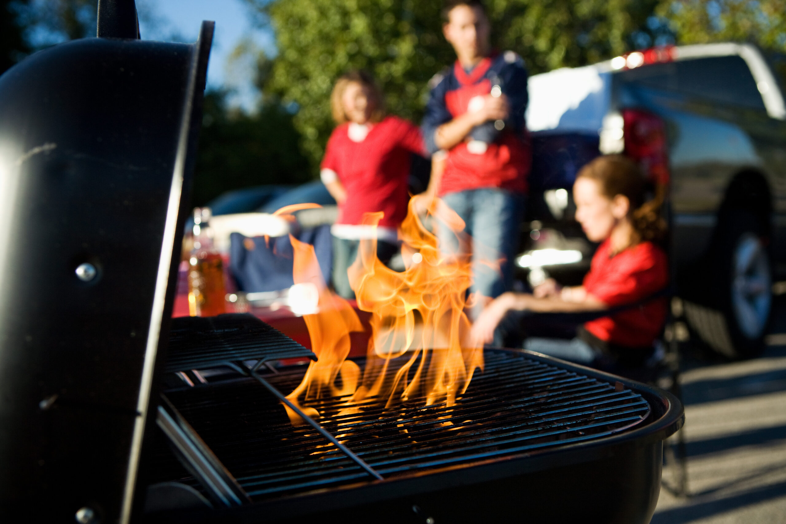 A family of three is in the middle of a tailgate party. The family is in the background and a flaming grill is in the foreground.