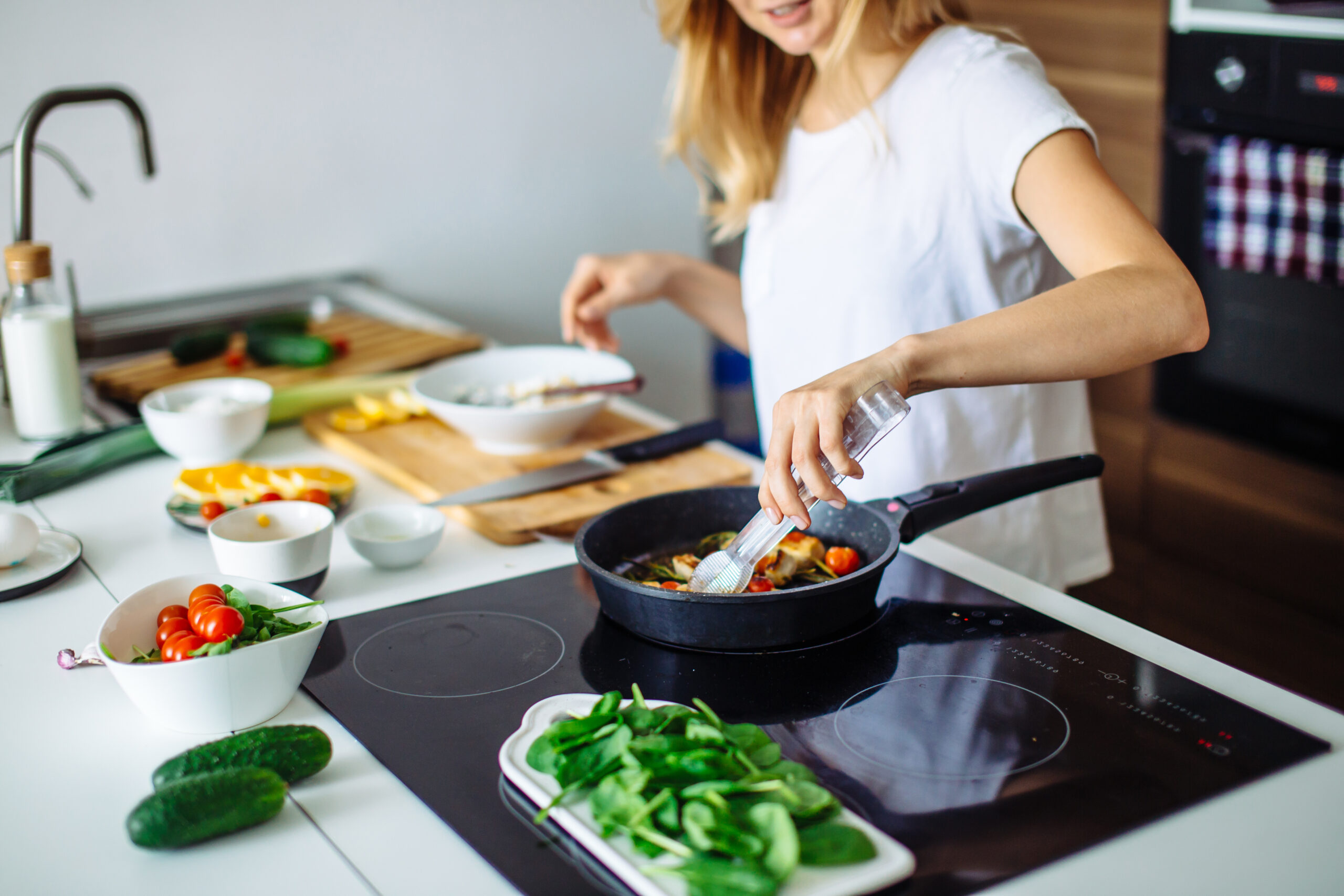 A young woman is using tongs to move vegetables around in a cast iron pan on a stovetop