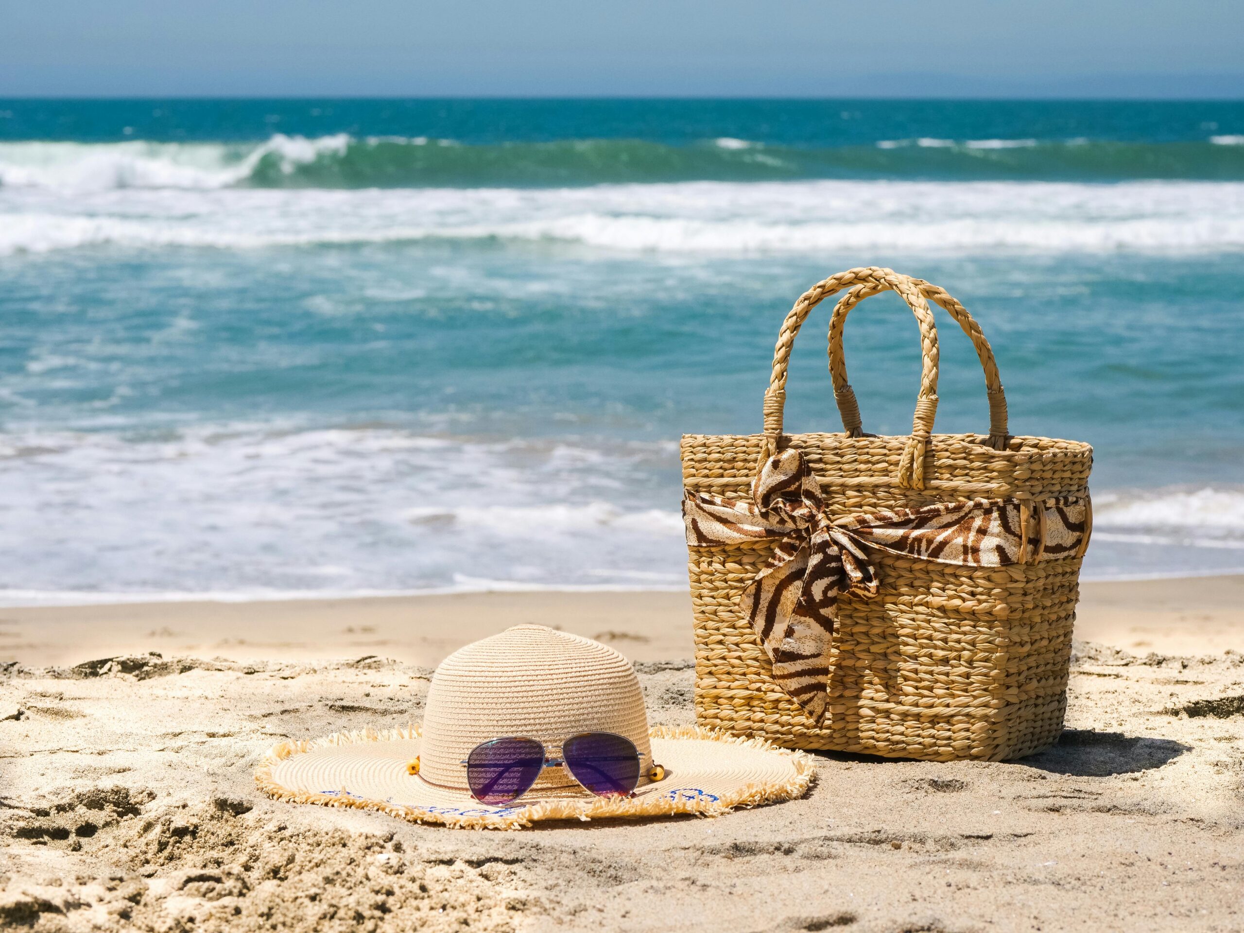 Beach hat, sunglasses and Beach Bag on the beach with ocean waves in the background.