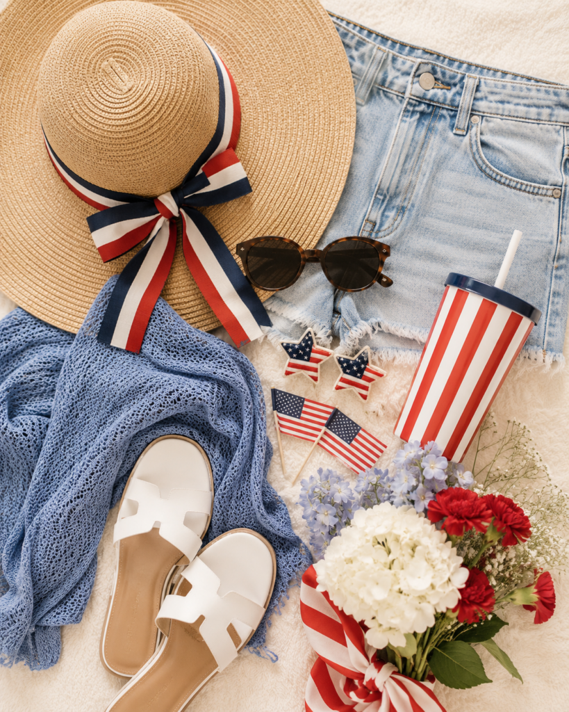 4th of July summer flat lay with a straw hat, denim shorts, sunglasses, white sandals, striped tumbler, mini American flags, and red, white, and blue flowers.