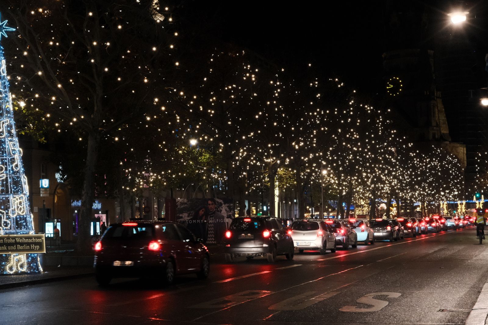 A downtown street lined with trees wrapped in Christmas lights, with cars driving through