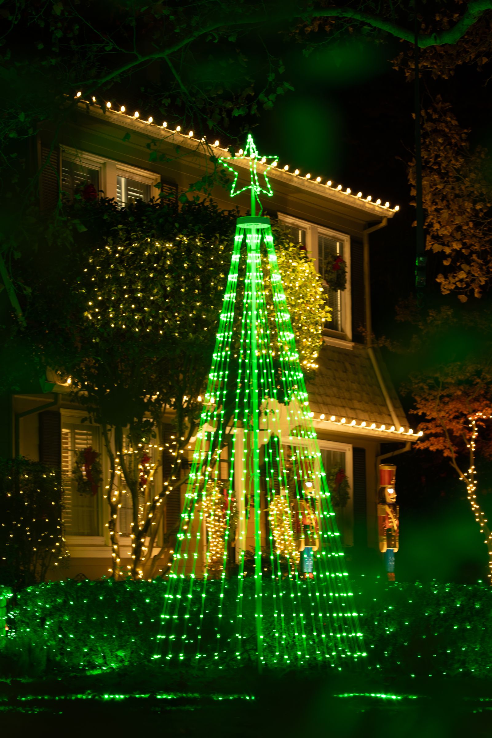 A house with a tall green light-up tree display in the front yard at night