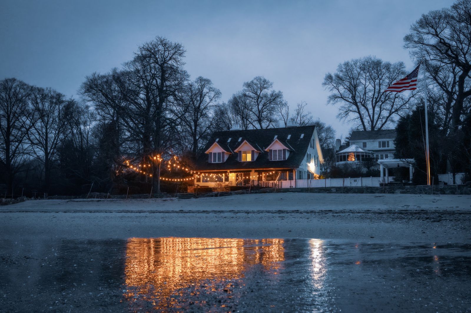 A lakefront house lit with Christmas lights reflecting on the water at dusk